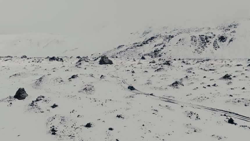 An aerial view shows a lone vehicle driving on a remote road through a vast, snow-covered, and desolate mountainous landscape