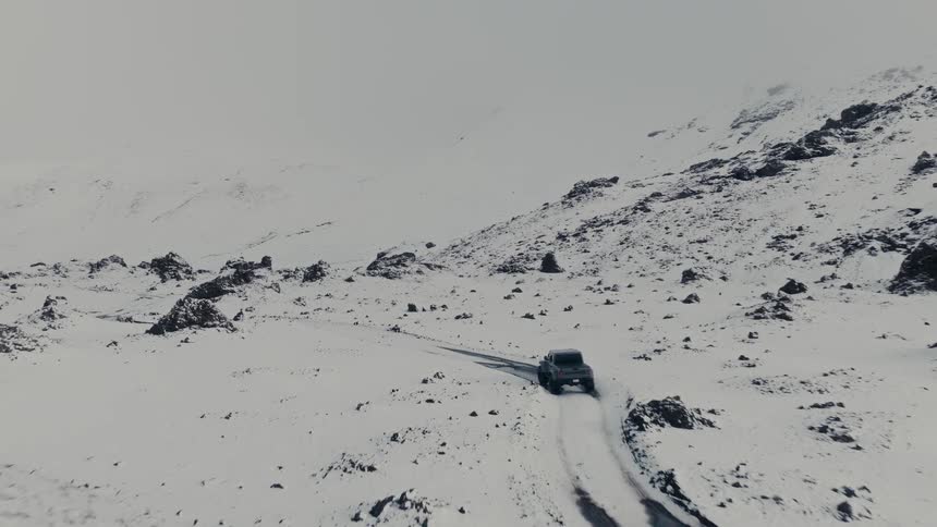 An aerial shot follows a rugged pickup truck driving through a vast, snow-covered mountain landscape