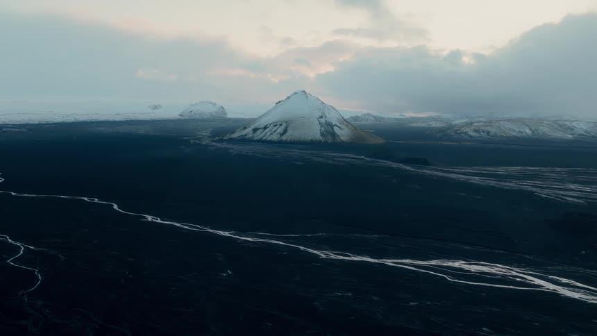 A vast and moody volcanic landscape features a snow-capped mountain rising above black sand plains and a winding river in Iceland