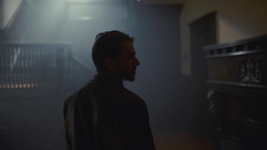A mysterious man stands in a dark, historic, and hazy room before turning to look at the camera