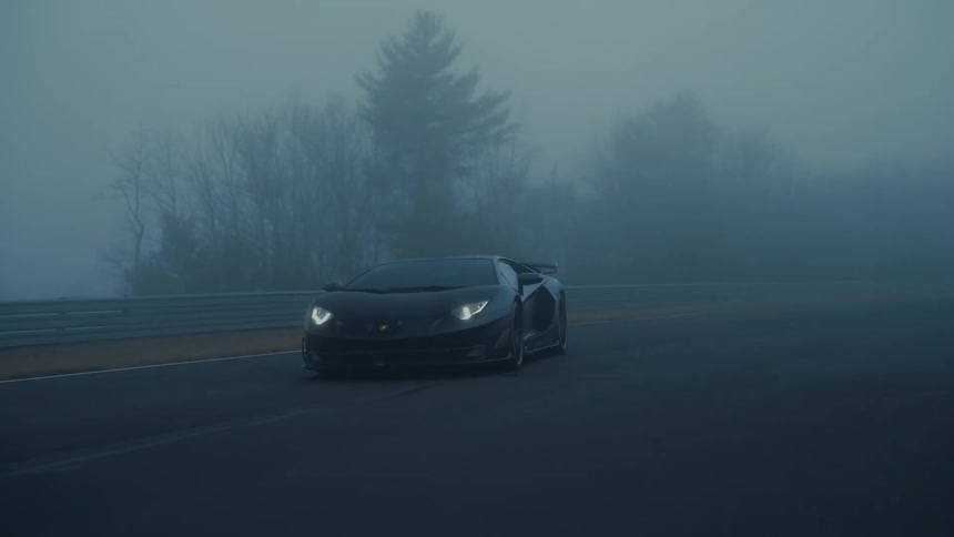 A sleek black Lamborghini with its headlights on emerges from the fog as it drives down a professional racetrack