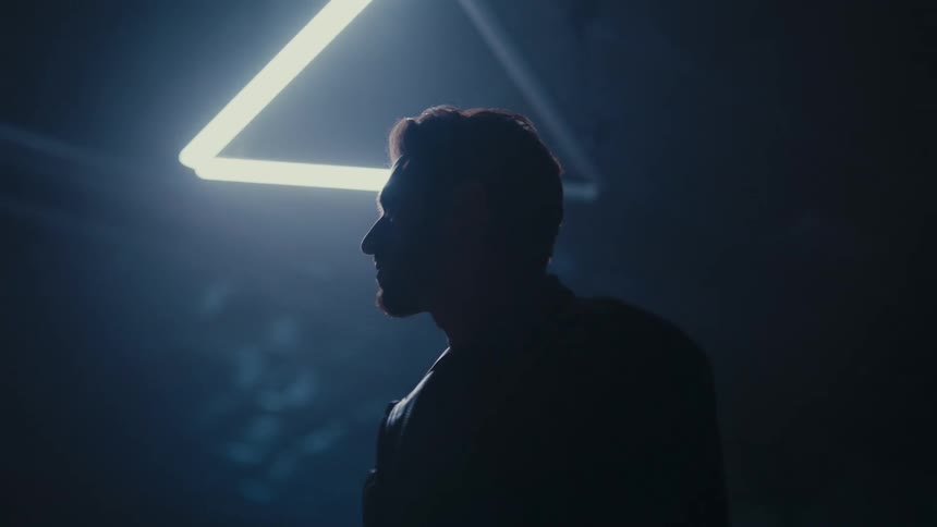 Cinematic shot of a man in a leather jacket turning slowly in a dark, smoky room beneath a triangular neon lamp