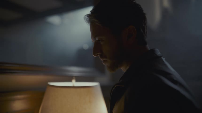 A somber man is illuminated by a single lamp in a dark, hazy room, appearing lost in his thoughts