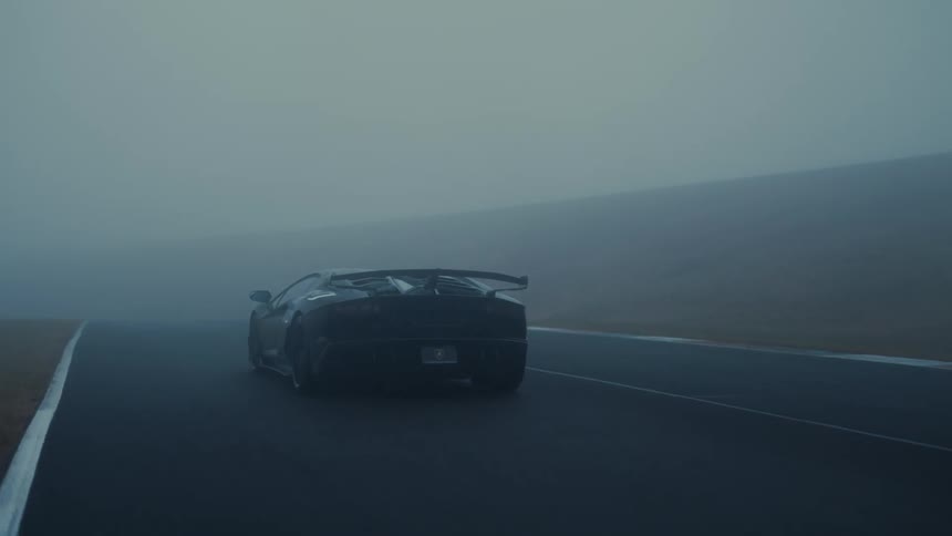 A powerful black supercar drives slowly along a winding racetrack enveloped in a thick, moody fog
