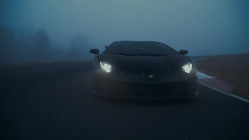 A sleek black Lamborghini with its headlights on is parked on a racetrack amidst dense, moody fog