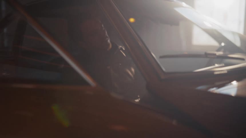 A man in a leather jacket gets into his vintage car in a moody, backlit garage