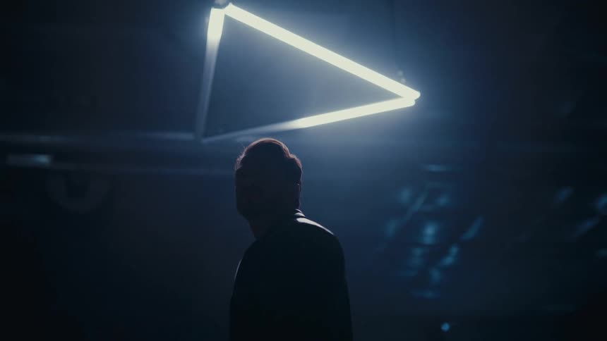 A mysterious man stands silhouetted under a glowing triangular light fixture in a dark, hazy room