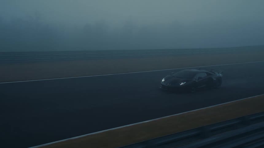 A sleek black supercar slowly emerges from the thick fog as it drives along a wet racetrack