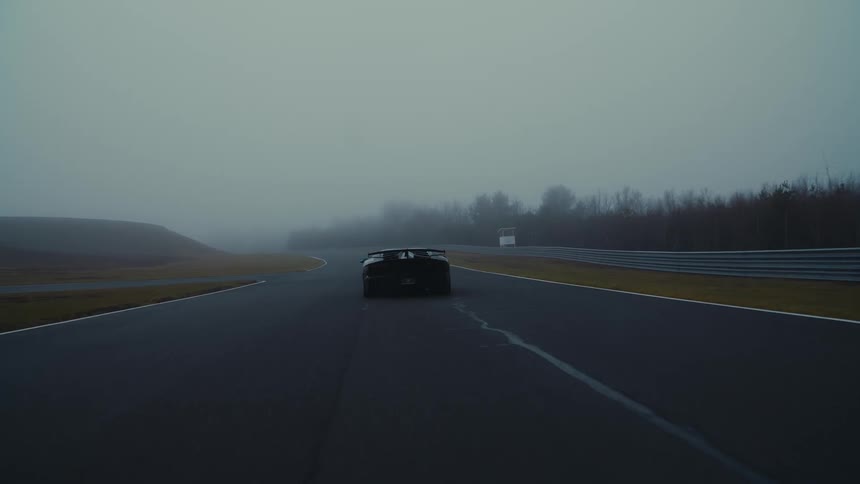 Drone shot pulls back to reveal a sleek black sports car driving away down an empty, foggy race track