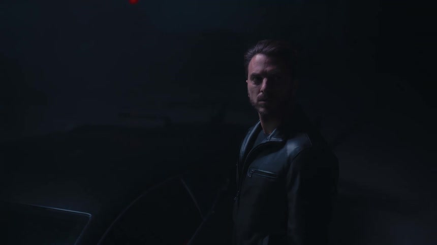 Slow zoom on a mysterious man in a black leather jacket standing in a dark, foggy garage with an intense stare