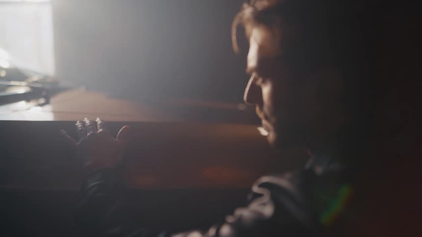 A pensive man in a leather jacket slowly caresses the hood of his vintage car in a dark, hazy garage
