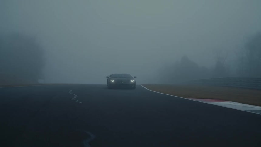 A sleek black luxury sports car slowly drives around a curve on a racetrack shrouded in dense fog