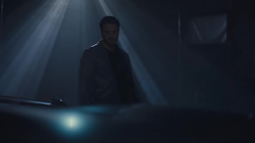 A mysterious man stands under a spotlight in a dark, hazy garage, staring intently forward