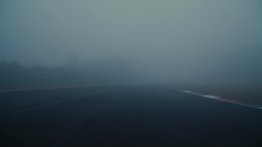A low-angle view shows a sleek, black sports car with its headlights on, driving fast towards the camera on a foggy racetrack