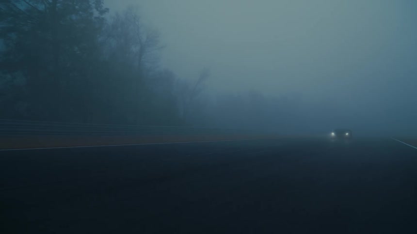 A sleek black Lamborghini with headlights on emerges slowly from the dense fog on a moody racetrack