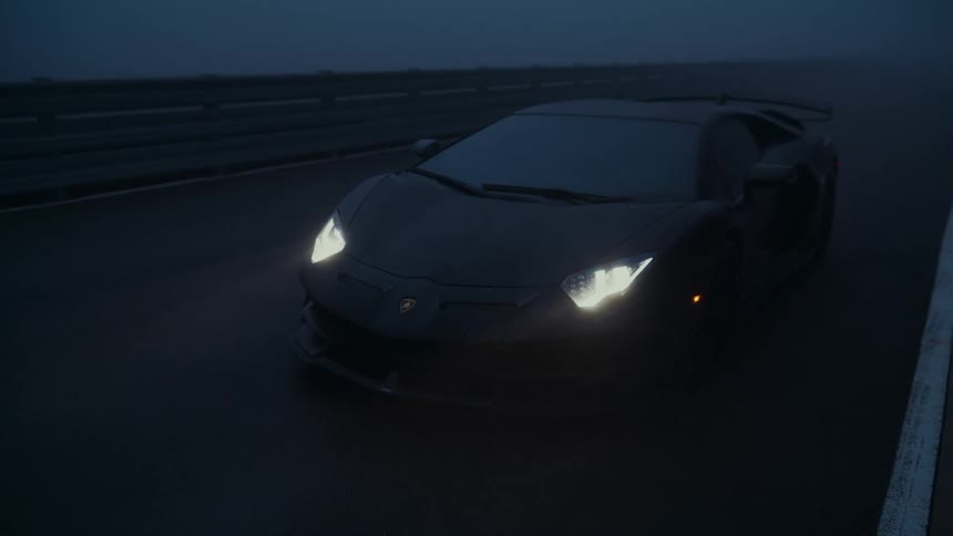Slowly pulling back from a matte black Lamborghini parked on a racetrack with its headlights cutting through the dense night fog