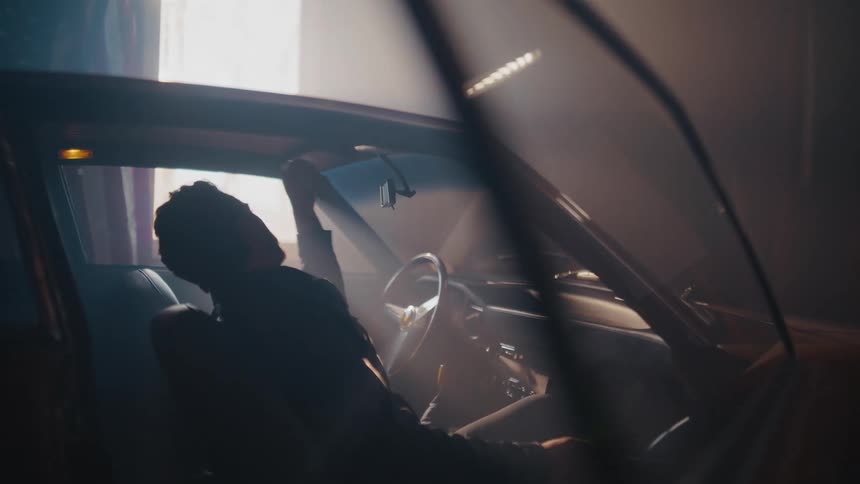 A man sits in the driver's seat of a classic muscle car in a dimly lit, hazy garage while using a tablet