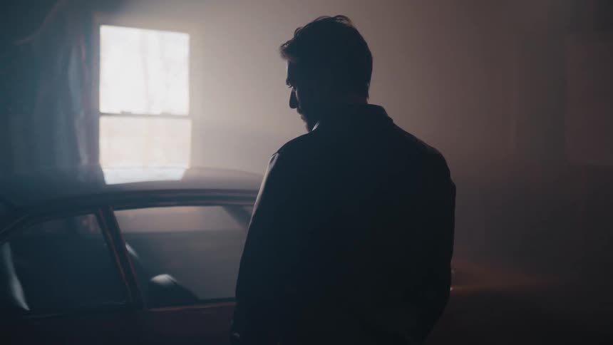 Pensive man silhouetted by a window stands next to his car in a dark and hazy garage