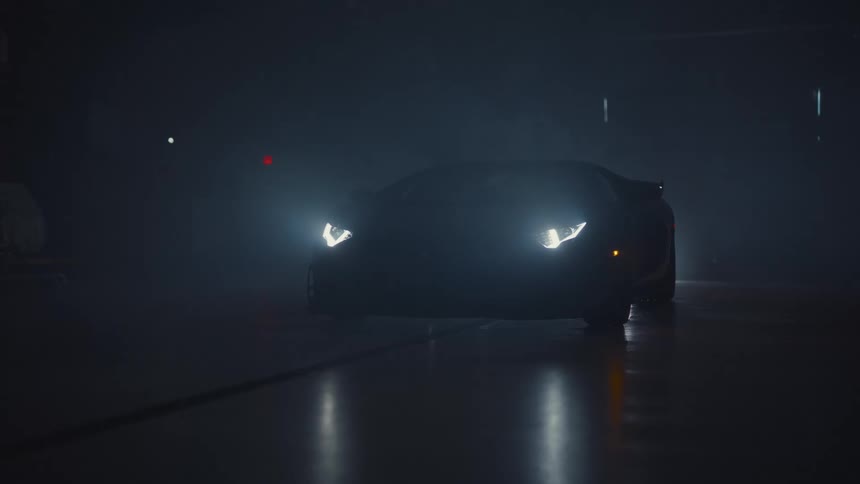 A black Lamborghini sports car turns on its bright headlights in a dark, foggy garage