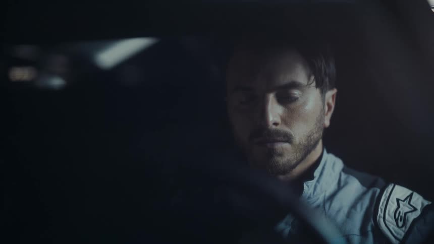 A focused male race car driver in a racing suit puts on his helmet inside his car at night