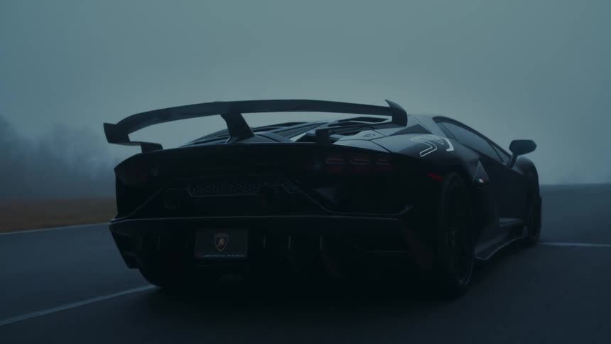 A sleek black Lamborghini glides around a racetrack enveloped in dense, atmospheric fog during the early morning