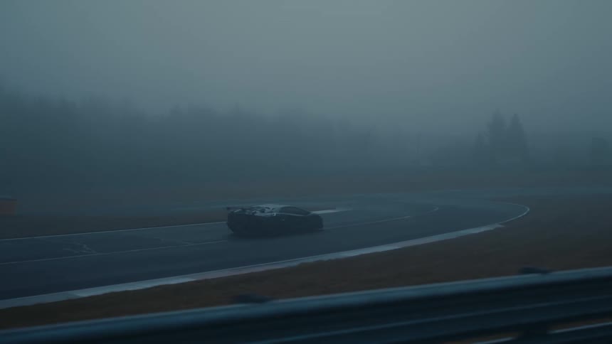 A sleek black supercar drives along a wet racetrack, disappearing into the thick, moody fog
