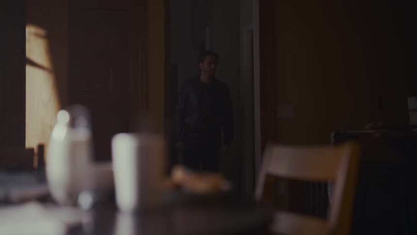 A mysterious man watches from a dark doorway as an untouched breakfast steams on the table in the foreground