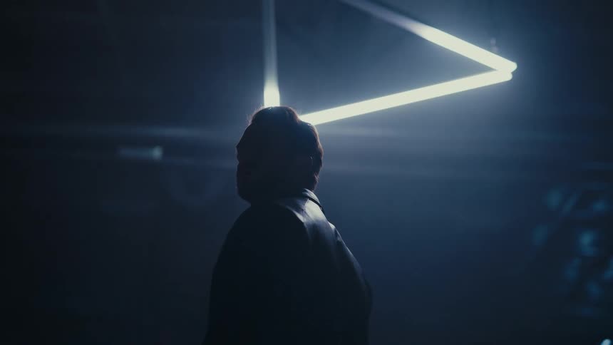 A woman's silhouette is cast by a bright triangular light in a dark, moody industrial space