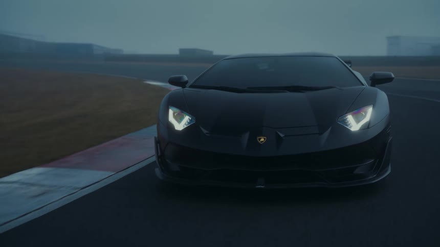 A sleek black supercar slowly navigates a turn on a wet racetrack enveloped in moody morning fog