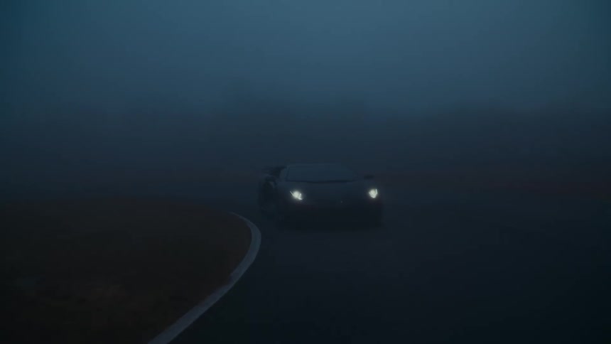 A sleek black sports car with bright headlights on drives slowly on a racetrack enveloped in thick, mysterious fog at dusk