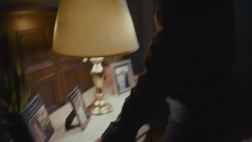 A somber man in a leather jacket gazes down at a pile of old, blood-stained photographs on a table in a dimly lit, smoky room
