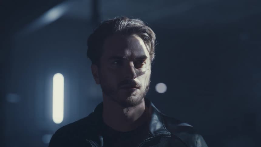 A mysterious man with a serious expression stands in a dark, hazy room as patterns of light and shadow move across his face