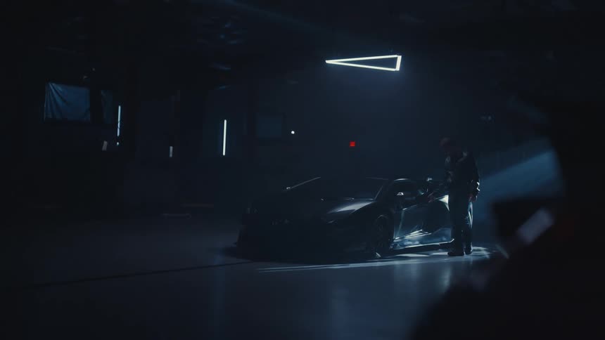 A man slowly approaches his luxury sports car parked inside a dark, hazy warehouse with dramatic spotlighting