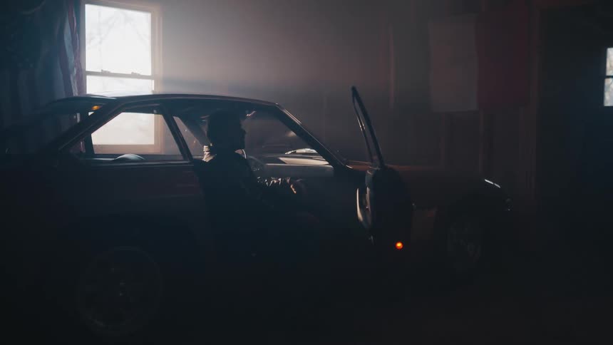 A silhouetted man gets into a classic sports car in a moody, hazy garage illuminated by a single window