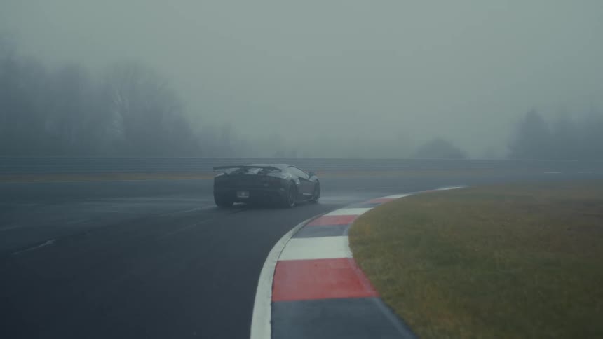 A sleek black Lamborghini supercar drives around a wet racetrack enveloped in dense fog