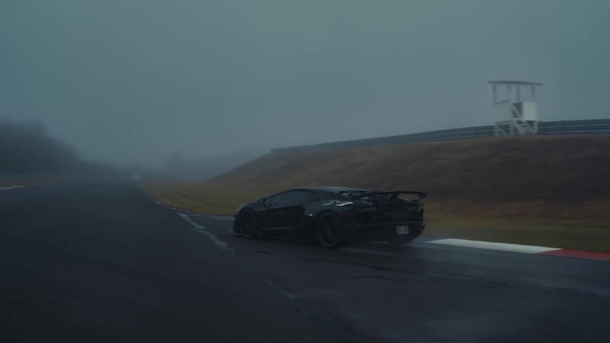A sleek black supercar navigates a winding racetrack, emerging from and disappearing into the dense, atmospheric fog on an overcast day