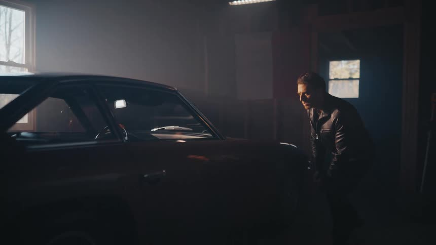 A man in a leather jacket inspects a vintage car in a dark, hazy garage while another person sits inside