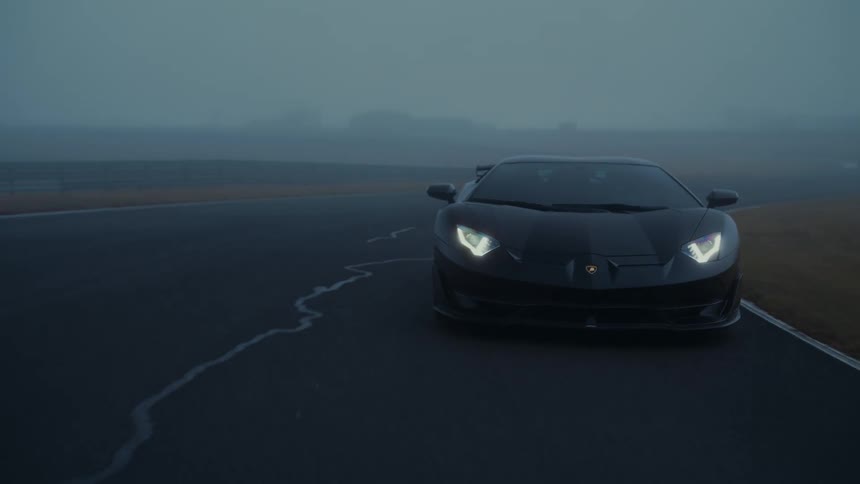 A sleek black Lamborghini with its headlights on slowly navigates a wet, foggy race track at dusk