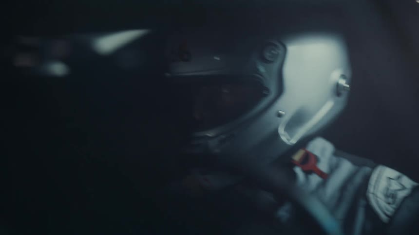 A focused professional race car driver sitting in a dark car removes his helmet and looks down