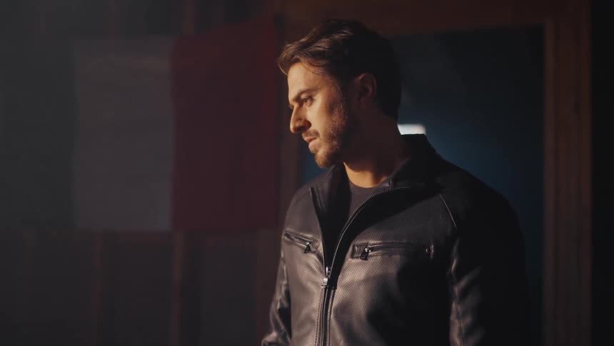 A serious man in a black leather jacket stands alone in a dark, smoky room, looking contemplative