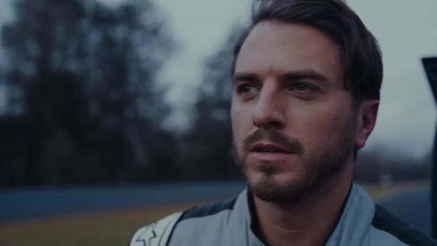 A determined male race car driver puts on his helmet next to a racetrack on an overcast day