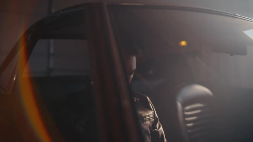 A moody man in a leather jacket sits thoughtfully in the driver's seat of his vintage car parked in a hazy, dimly lit garage
