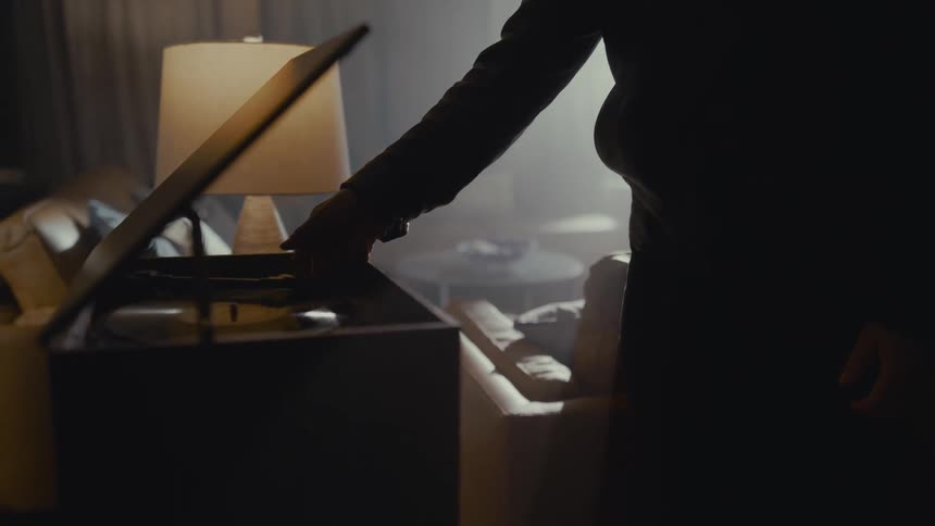 A silhouetted man in a dimly lit, atmospheric room carefully places the needle on a vintage record player