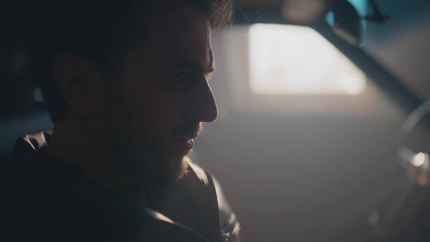A serious man sits in the driver's seat of a car in a dark, hazy garage, looking down thoughtfully