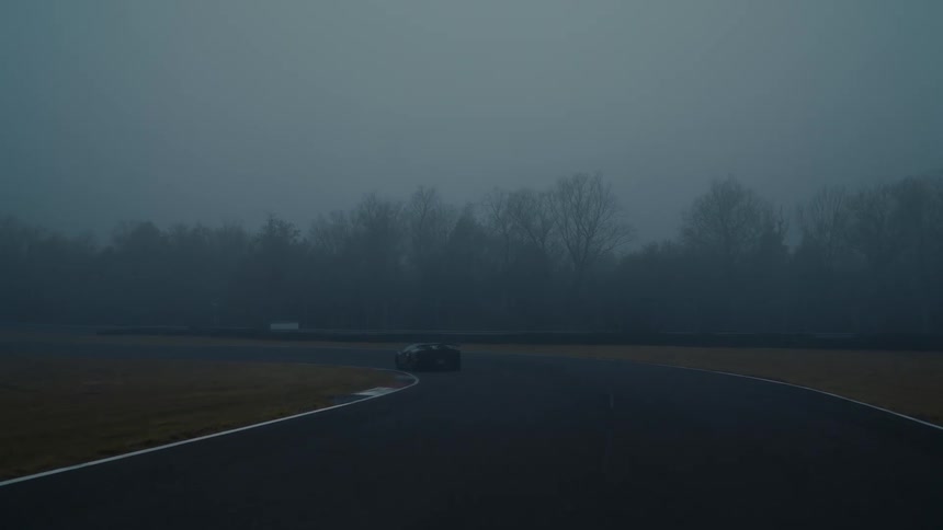 A sleek sports car drives along a wet, winding racetrack on a moody and foggy morning