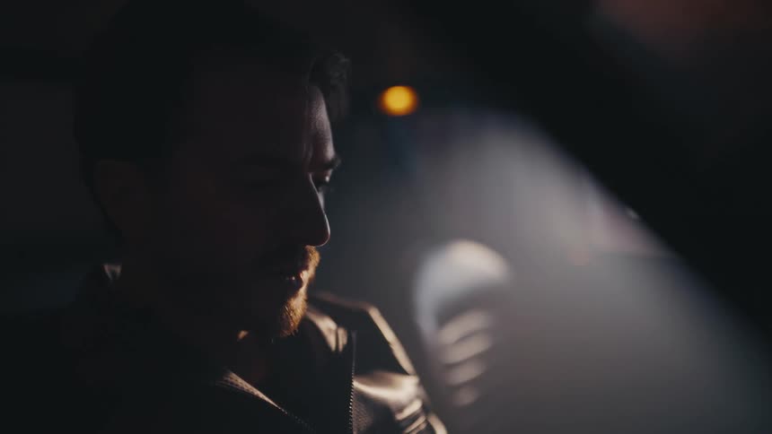 A pensive man wearing a leather jacket sits alone in his car at night, dramatically lit and deep in thought