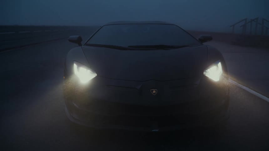 A sleek black Lamborghini sits on a wet road in the dark, its headlights cutting through the dense fog and rain