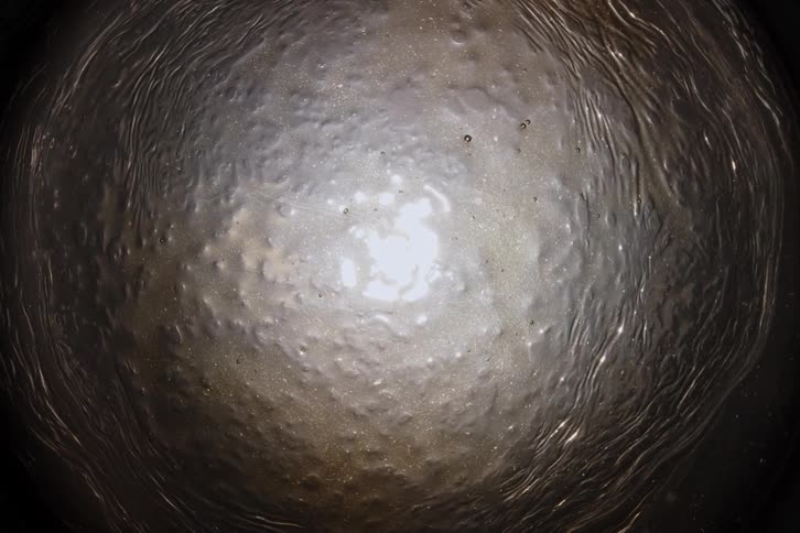 A mesmerizing top-down, abstract view of a thick, silvery liquid slowly undulating inside a dark, circular container