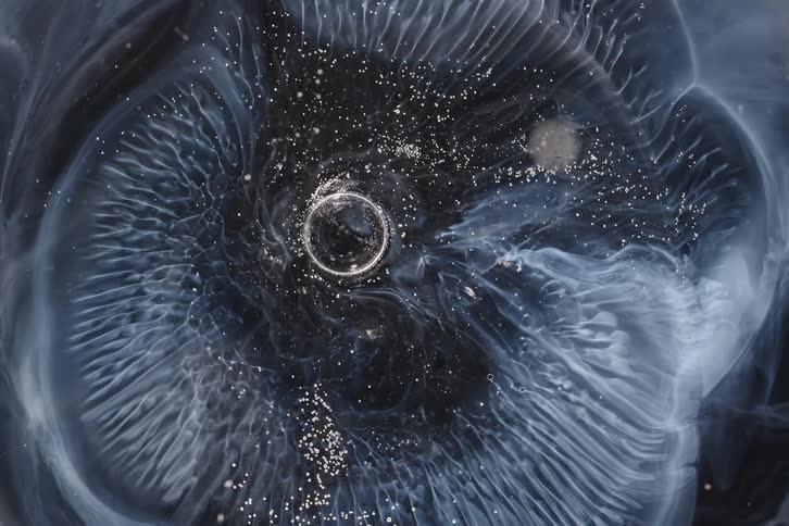 A mesmerizing cosmic vortex of swirling white ethereal liquid and sparkling particles slowly expands against a dark background