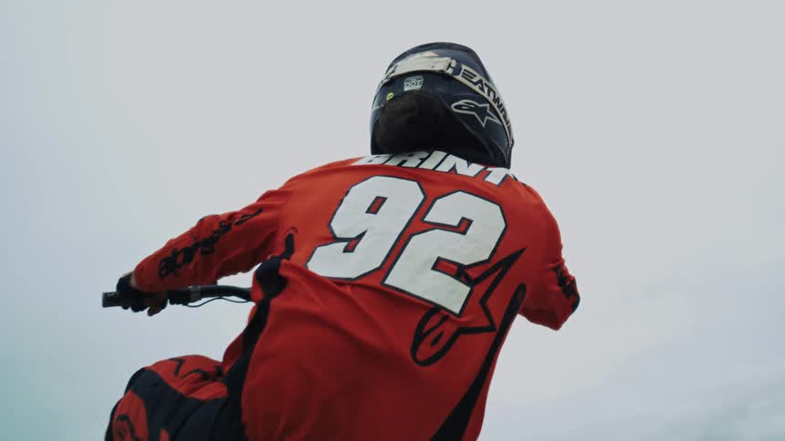 A low-angle slow-motion shot from behind a motocross rider in red racing gear looking up a sand dune